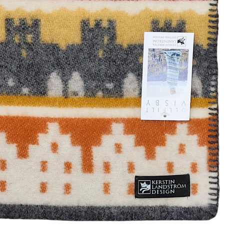 A Swedish blanket made from Norwegian lamb’s wool, featuring orange, yellow, and gray geometric patterns, a black Kerstin Landström Design label, and a product tag underneath.