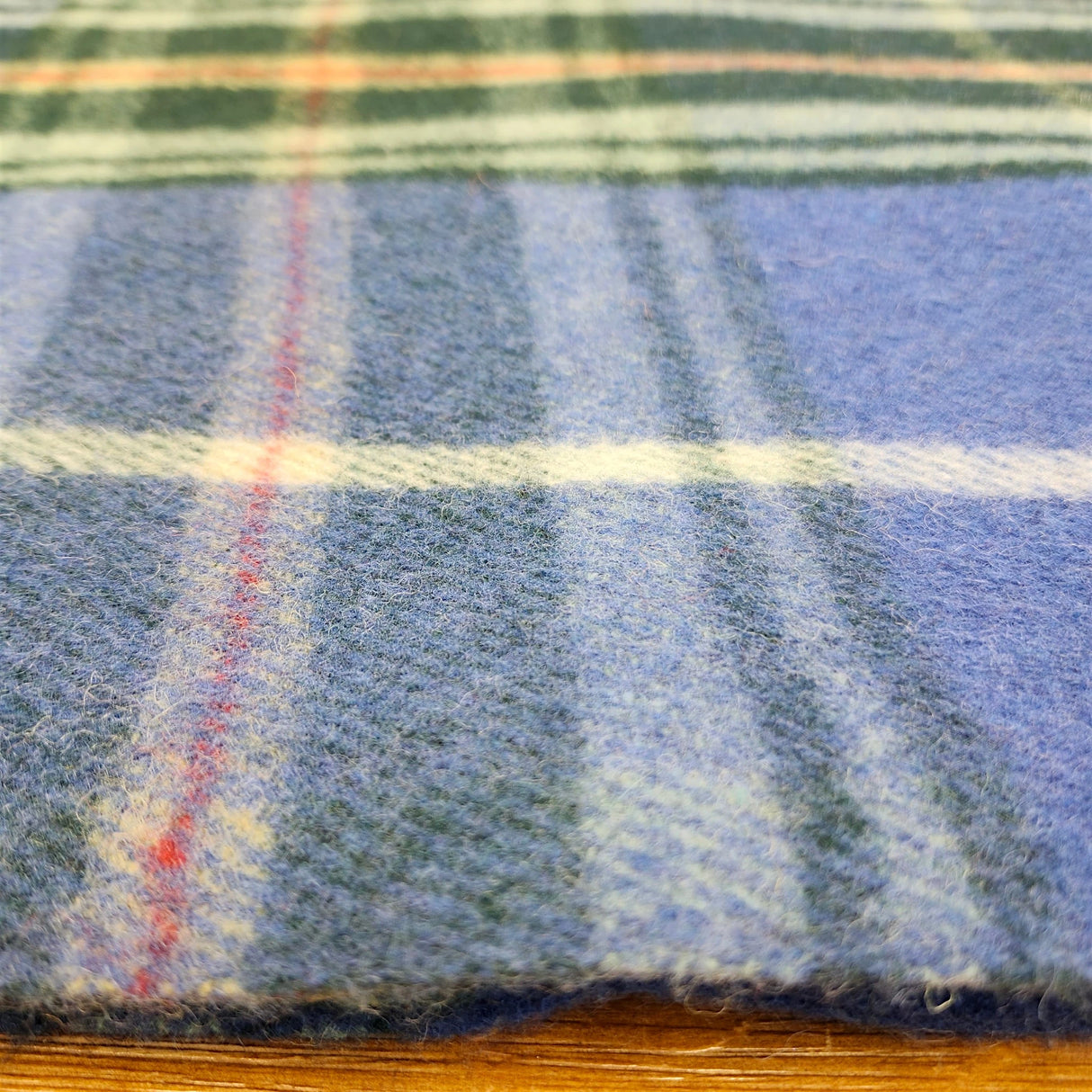 Highland tartan blanket in blue, white, green, and red plaid displayed flat on a wooden surface, showing its check pattern and soft woven texture.