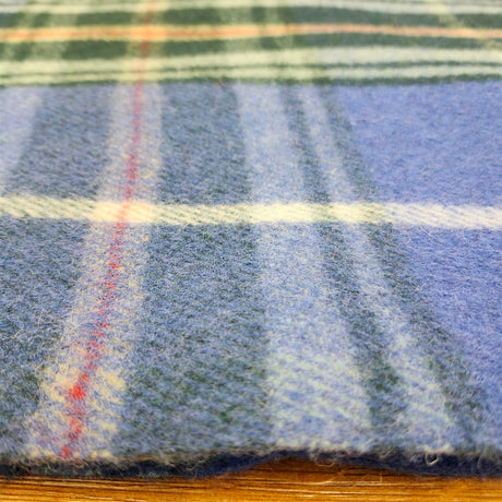Highland tartan blanket in blue, white, green, and red plaid displayed flat on a wooden surface, showing its check pattern and soft woven texture.