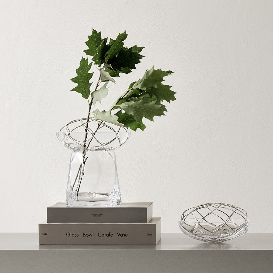 The Orrefors: Bloom Vase Low with a geometric design holds leafy branches atop stacked books, while a matching crystal bowl rests nearby on a minimalist surface.