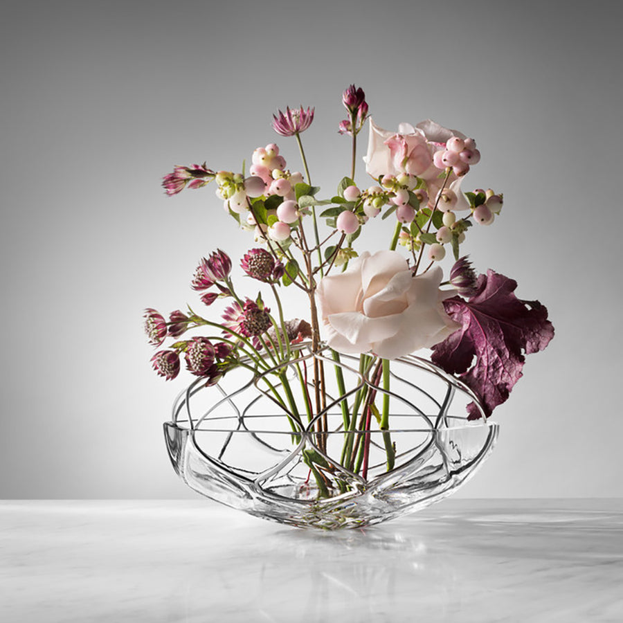 The Orrefors: Bloom Vase Low showcases a lovely white and pink flower arrangement with dark purple leaves against a gray background on a white surface.