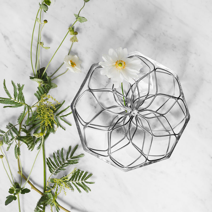 The Orrefors Bloom Vase Low, a hexagonal glass piece with a metal grid, holds a single white flower, complemented by greenery on a marble surface.