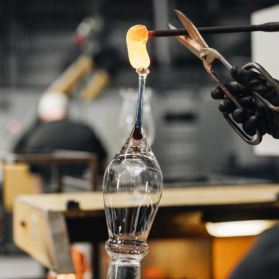 In a workshop reminiscent of Orrefors, a glassworker skillfully shapes hot glass with precision tools, evoking the timeless elegance seen in the Intermezzo Blue Aroma Glass 20oz by Erika Lagerbielke.