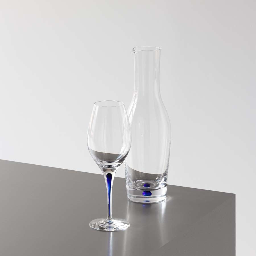 A mouth-blown crystal wine glass and carafe with a blue dot detail, from the Orrefors Intermezzo Blue Balance (2-Pack), gracefully sit on a gray surface against a plain white backdrop.