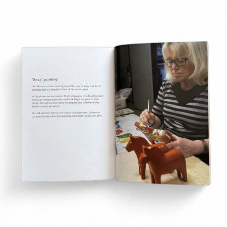 A blonde woman with glasses paints two small wooden Dala Horses at a table, while the open book "The Dala Horse, History Tradition Handicraft by Swedish Icons Paperback" lies nearby, showing text and images on Swedish icons.