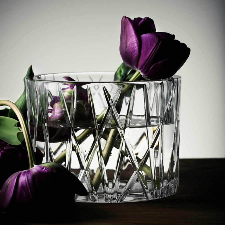 An Orrefors City Bowl filled with water showcases vibrant purple tulips on a wooden surface, with one gracefully bent over the table.