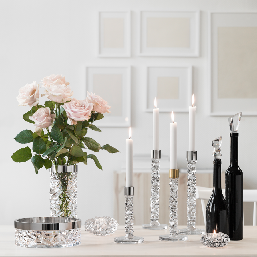 A stylish table setting features crystal candle holders with lit candles, a Carat Bowl by Orrefors, and a vase of pink roses. Black bottles and décor are arranged on a white table against a light wall adorned with framed art by Lena Bergström.