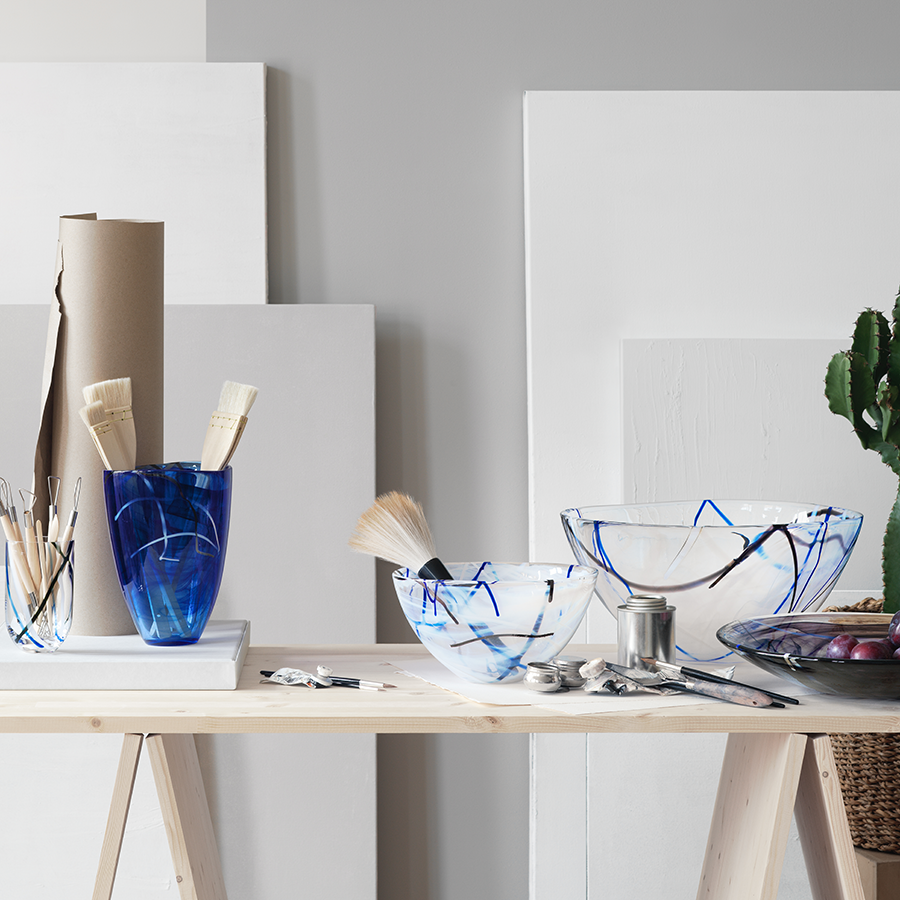 A wooden table features paintbrushes in a blue vase, paired with ceramic bowls similar to KostaBodas Contrast Blue Bowl Medium. Assorted utensils enhance the display, with rolls of paper and panels subtly in the background.