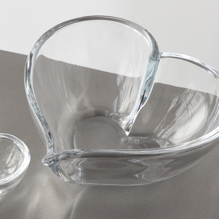 An Orrefors Heart Bowl Large, a transparent heart-shaped glass piece, elegantly rests on a gray surface beside a decorative small round glass dish.