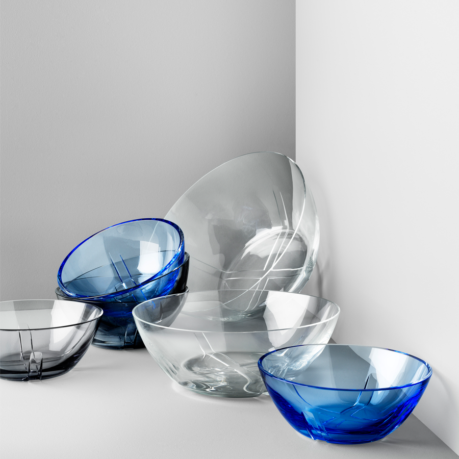 A collection of clear and Kosta Boda blue glass bowls, featuring the elegant Bruk Bowl Blue Small from the KostaBoda range, are arranged against a white and gray corner wall.