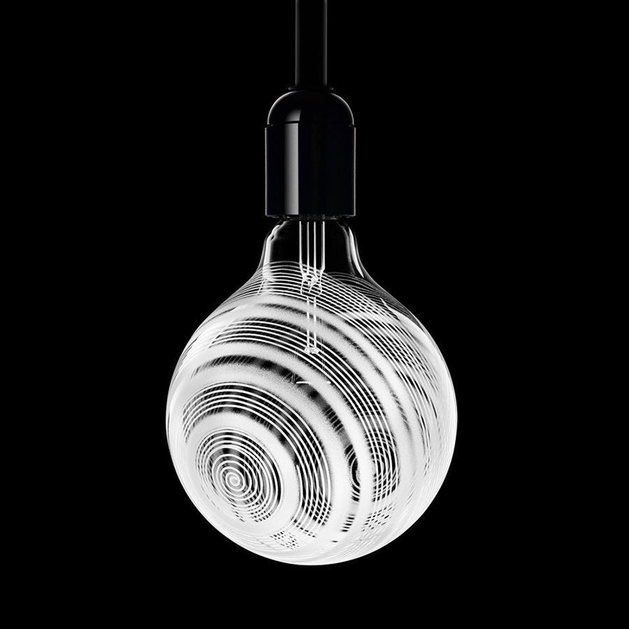 The Orrefors Light Shadow Bulb Loop 25W, set against a black background and mounted on a sleek black fixture, captures minimalist design with its decorative spiral patterns.