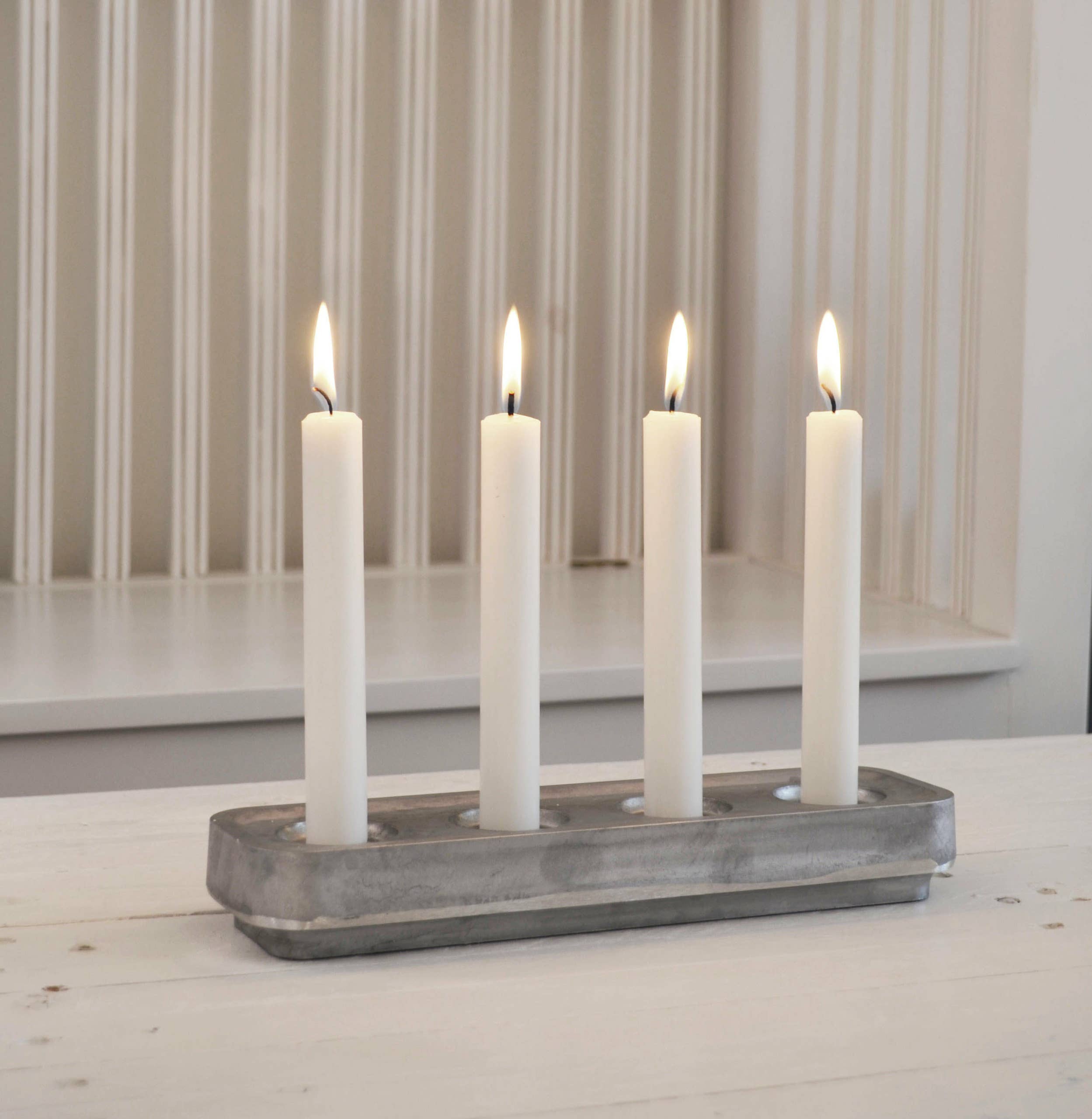 Four lit white candles sit in a Stumpastaken Black Powder Coat Swedish Candle Holder by Jonas Torstensson on a white wooden surface with vertical grooves in the background.