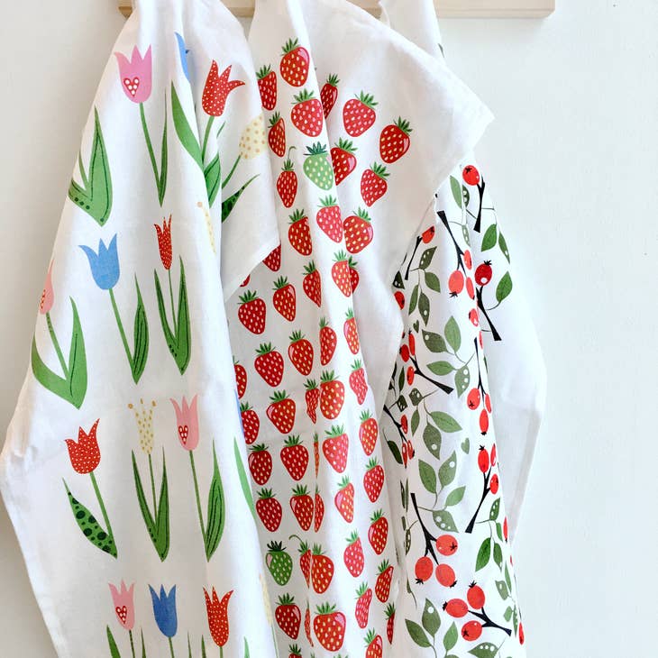 A gift duo featuring an ecofriendly dishcloth with a tulip pattern, along with additional dishcloths adorned with strawberries and cherries, hang on a white wall.