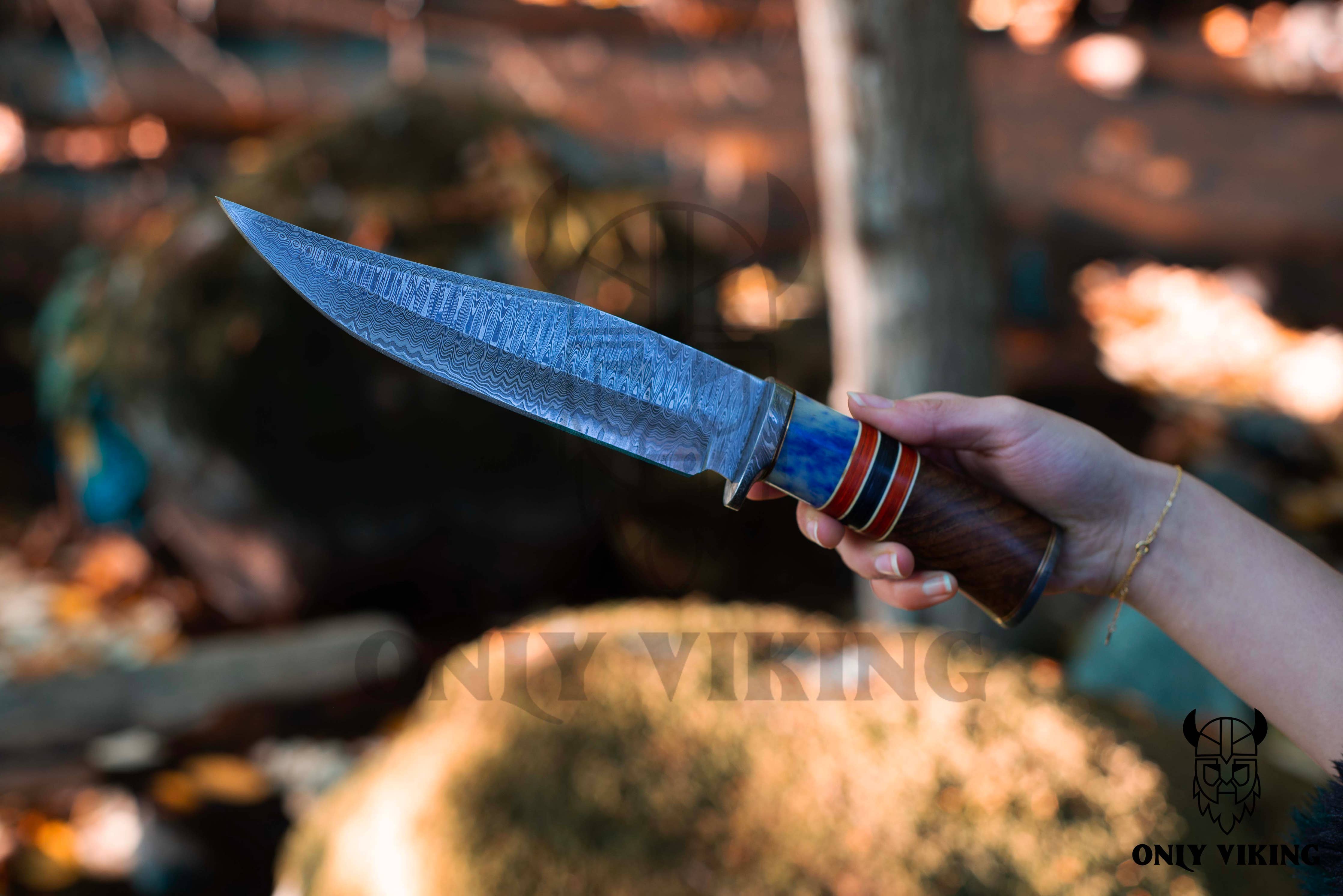 A hand holds the Damascus Steel Hunting Knife, featuring a patterned blade and a vibrant handle, set against an outdoor wooded backdrop that highlights its rugged appeal.