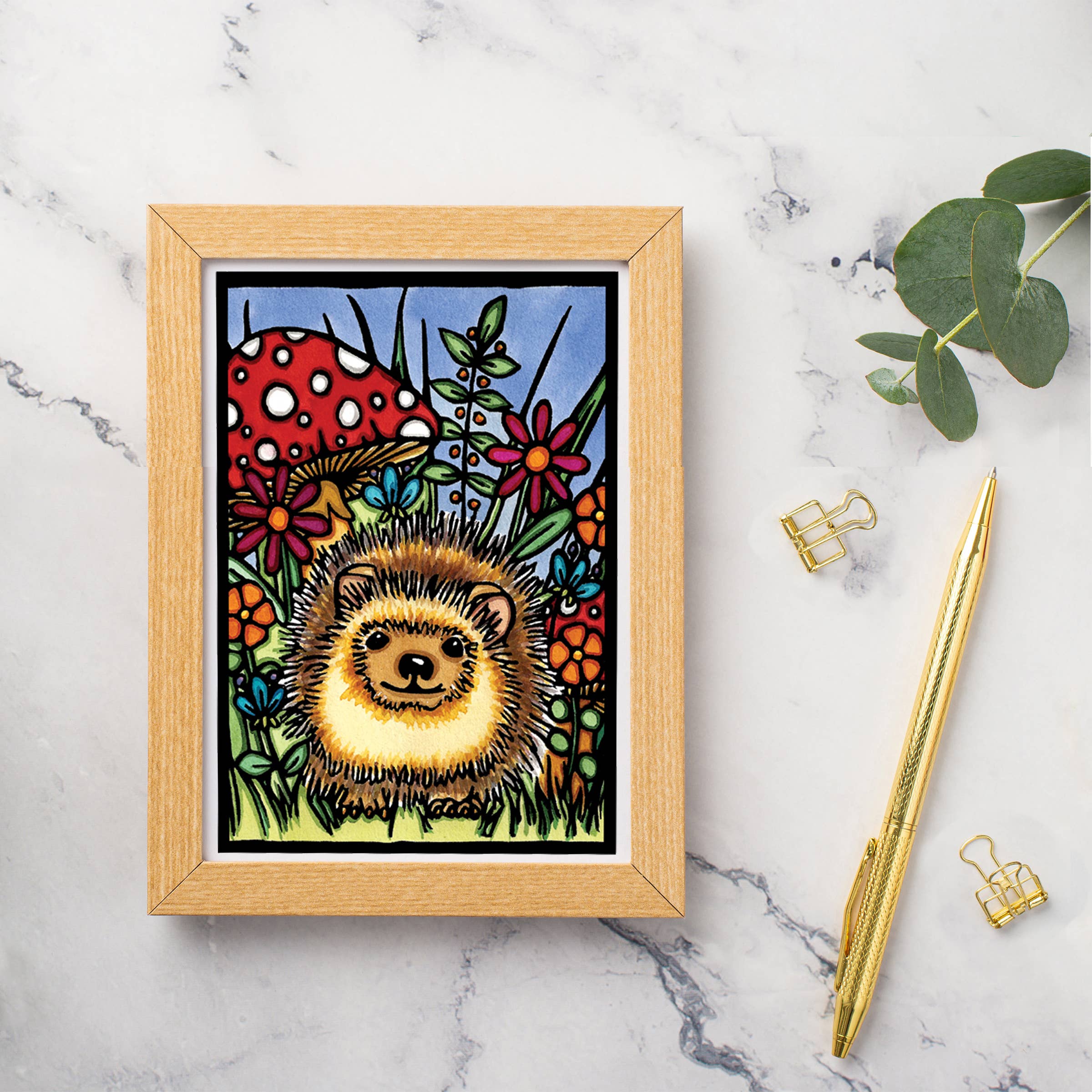 A greeting card titled "Hedgehog" by Sarah Angst, featuring a hedgehog surrounded by colorful flowers and a red mushroom, is placed on a marble surface next to a gold pen, two clips, and eucalyptus leaves.