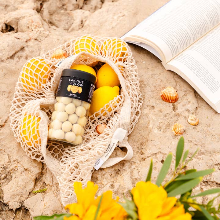 A mesh bag with lemons and a jar of Lakrids by Bülow Lemon liquorice candies sits on a rocky surface, nestled beside an open book, seashells, and vibrant yellow flowers.