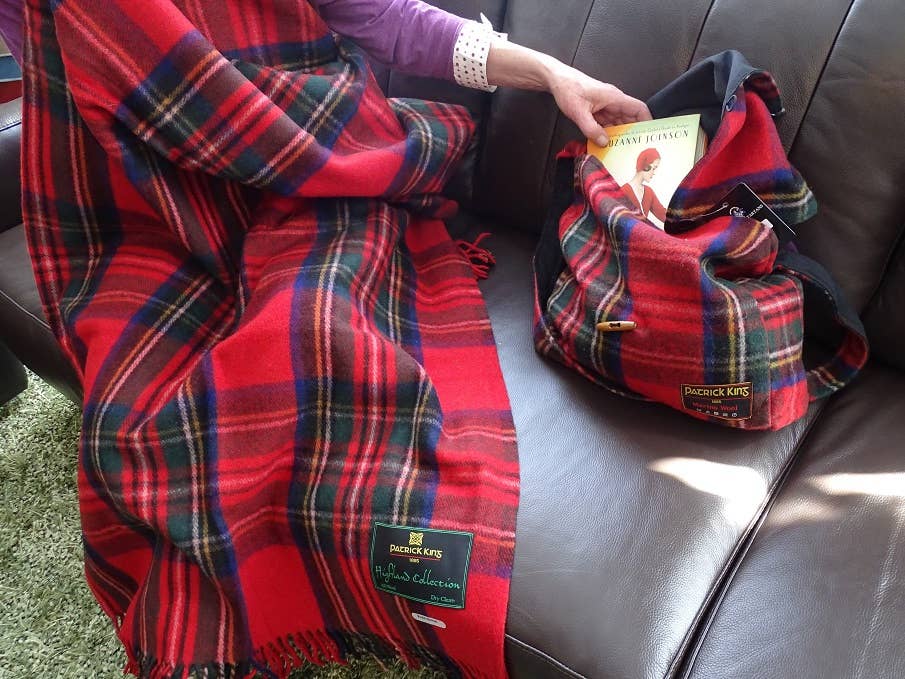 A person on a brown sofa holds a book and a tartan bag that matches the Royal Stewart Deluxe Highland Blanket draped over their lap.
