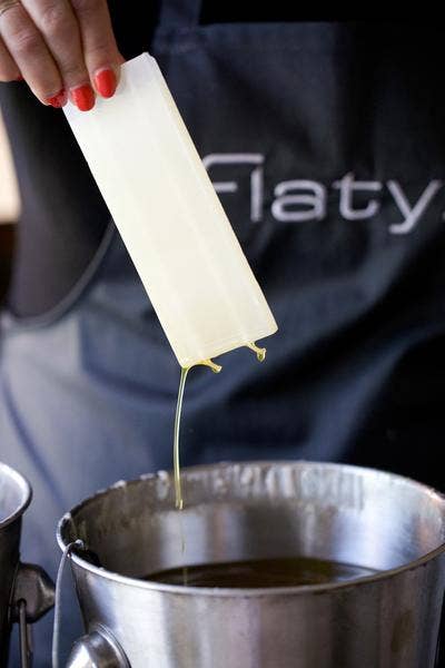 A hand holds a wax strip with melted wax dripping over a metal pot; the person, in a dark apron, prepares to craft a Candle: Wine in Vineyard - Flat Candle Flatyz.