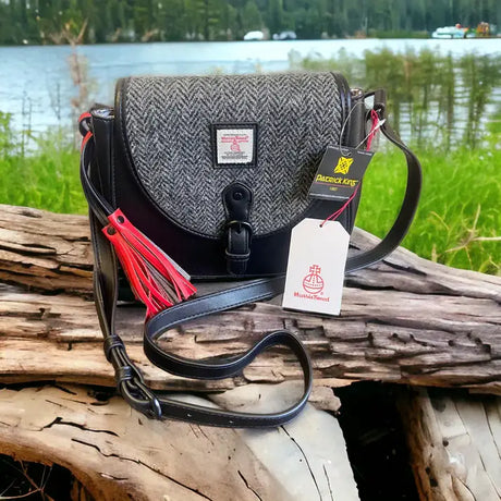 The Harris Tweed Saddle Bag - Grey, featuring a herringbone pattern and red tassel, rests on a log with a scenic lake and trees behind it. Two tags are attached to the bag.