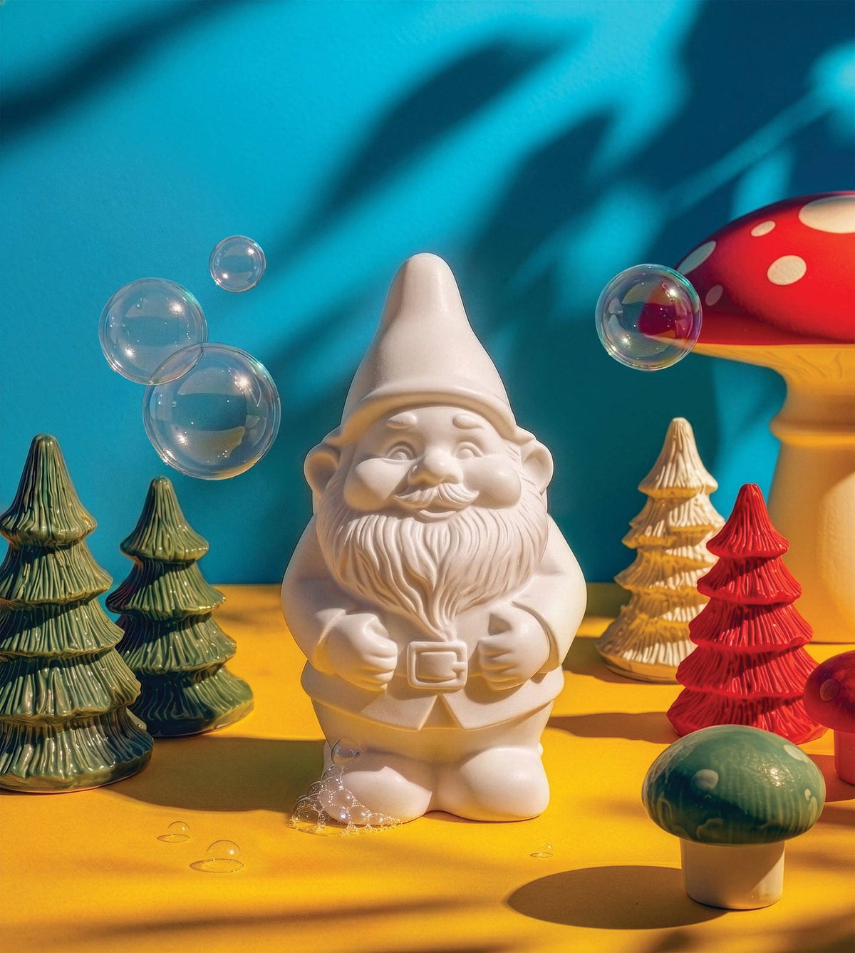A white garden gnome with ceramic trees, mushrooms, soap bubbles, and a Swedish Pine soap bar arranged on yellow, set against a blue background.