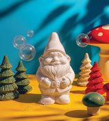 A white garden gnome with ceramic trees, mushrooms, soap bubbles, and a Swedish Pine soap bar arranged on yellow, set against a blue background.