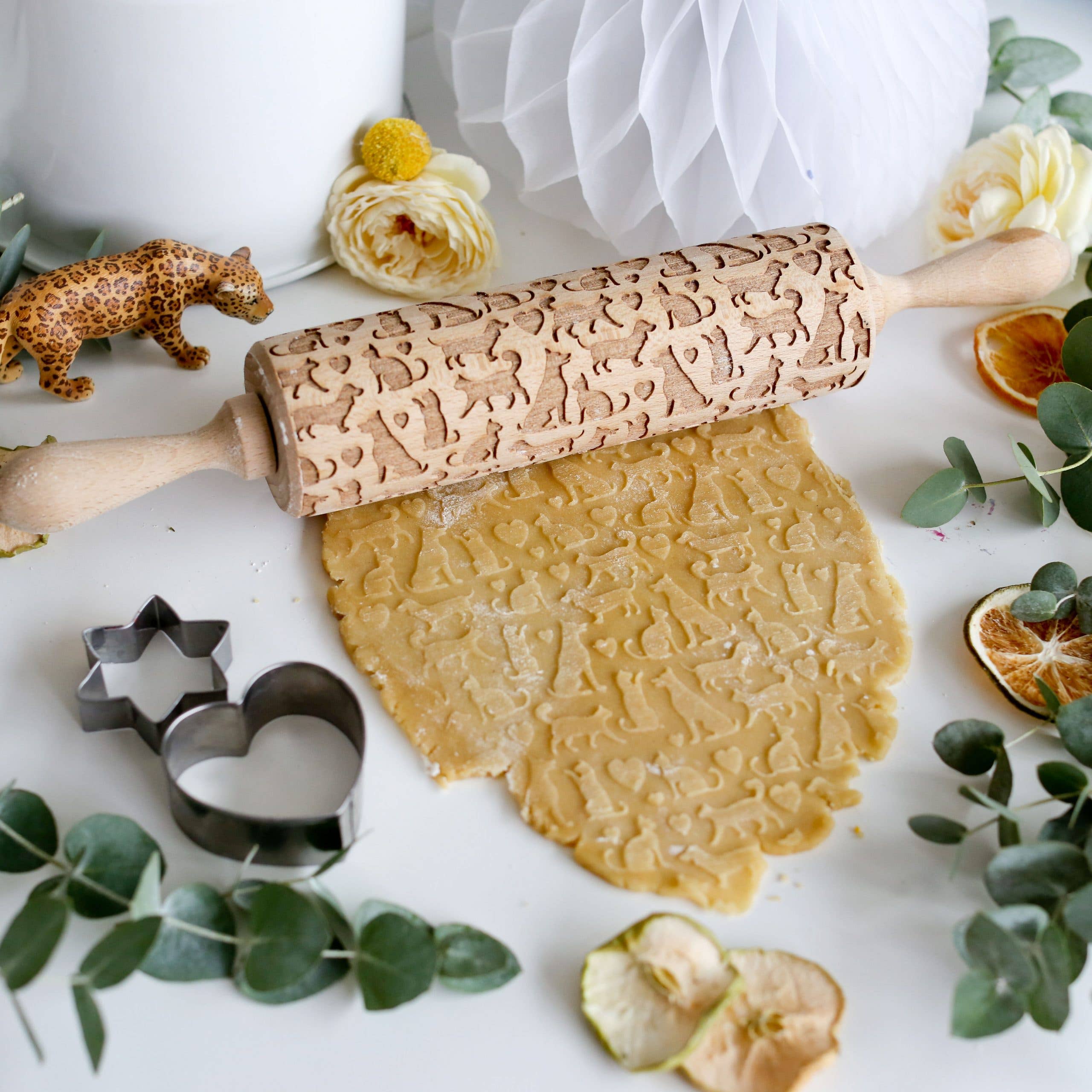 The Pet Love Embossing Rolling Pin with Cats & Dogs creates charming animal patterns on dough, surrounded by cookie cutters, eucalyptus leaves, dried oranges, yellow flowers, and a small toy leopard.