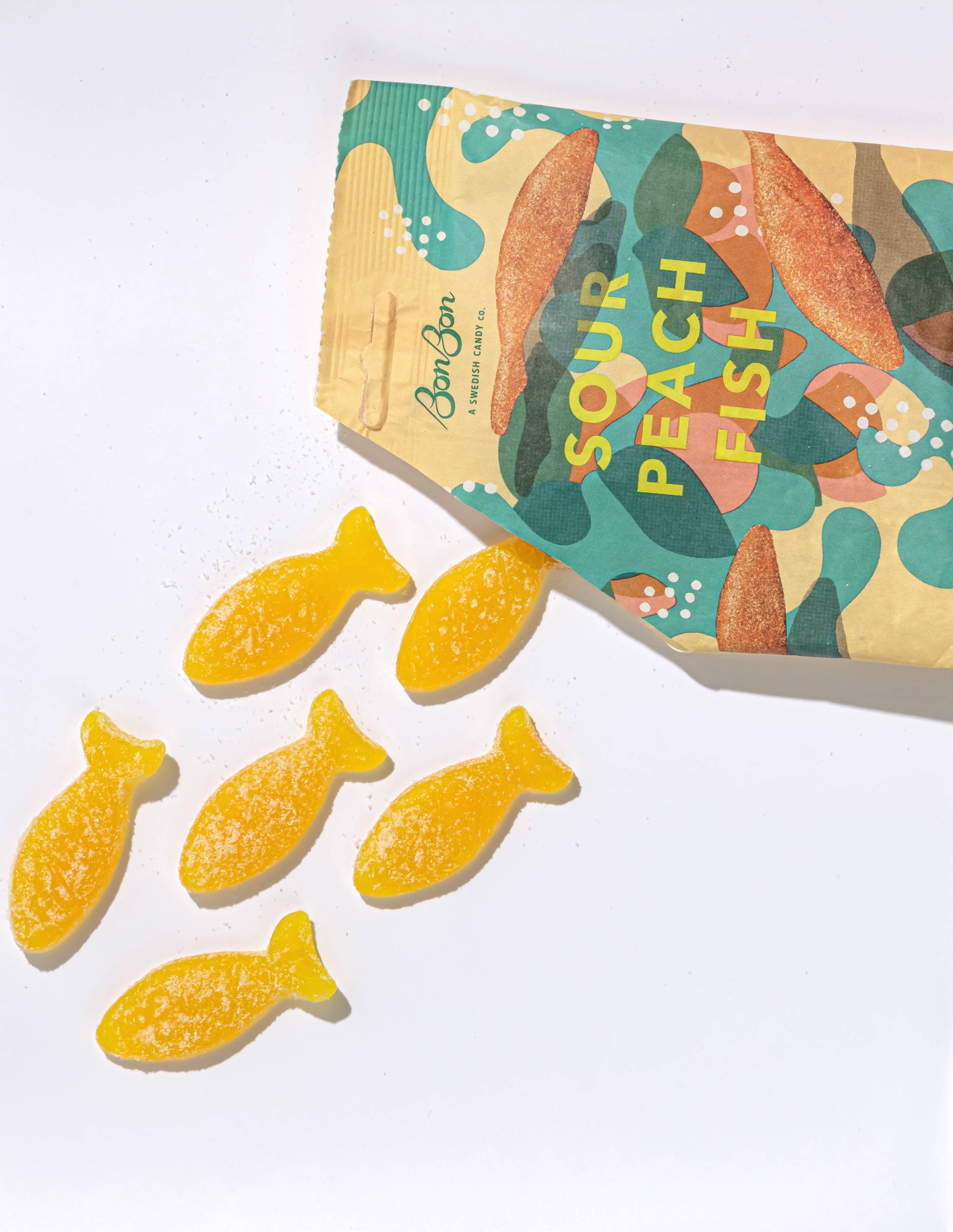 An open bag of Candy: BonBon - Sour Peach Fish (150g) Swedish Candy reveals six yellow, vegan gummy candies shaped like fish scattered around it.