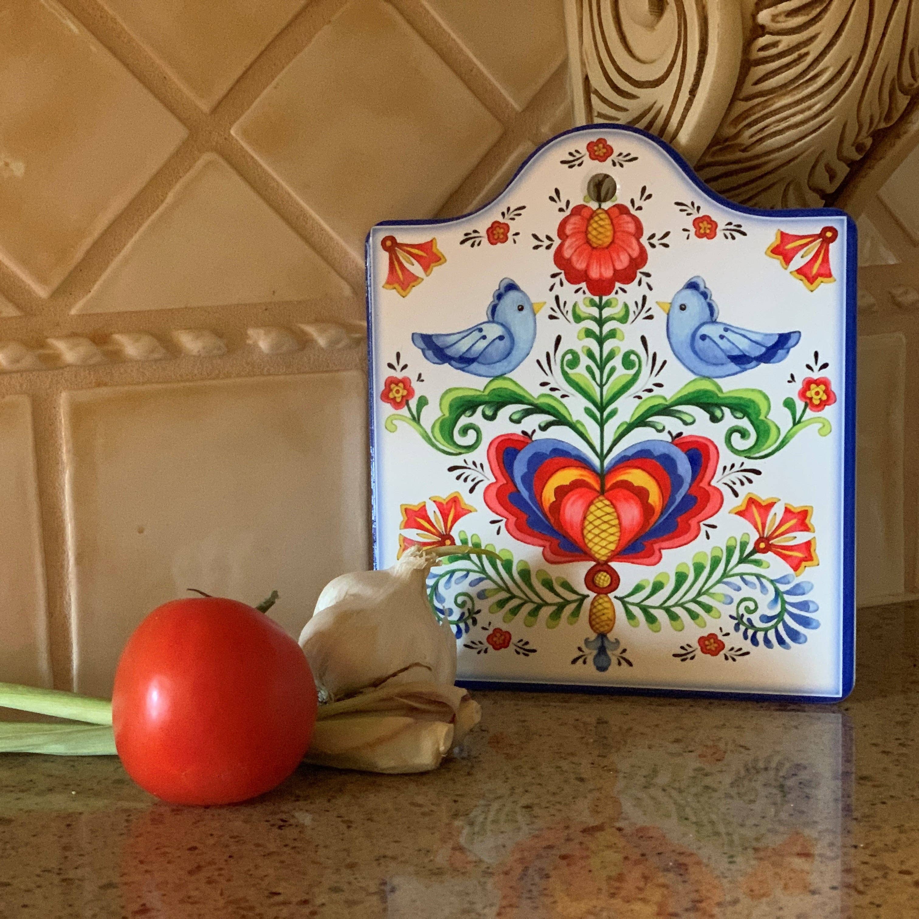 The Lovebirds Folk Art Cork Backed Ceramic Cheeseboard, featuring a floral and lovebirds design, sits on a kitchen countertop beside a tomato, garlic, and green onion, infusing the space with Scandinavian charm.