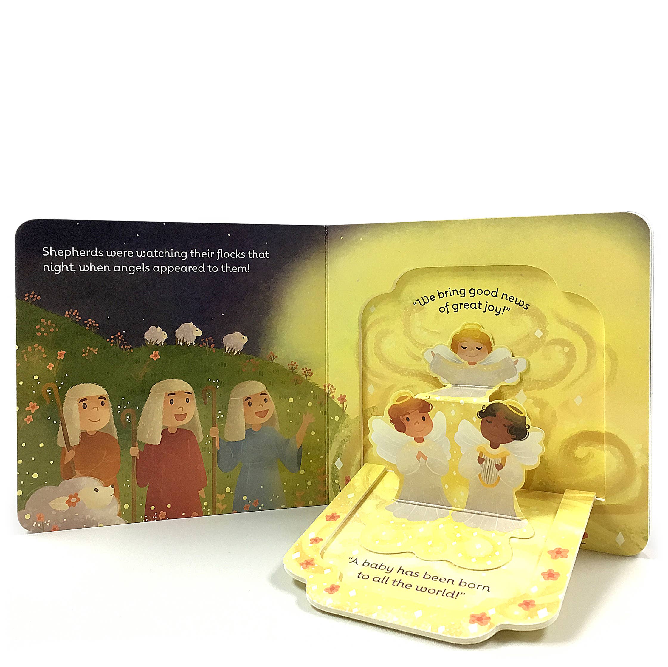 An open Book: First Christmas Lift-a-Flap Baby Jesus Board Book shows shepherds and sheep on one page, and three angels with Baby Jesus plus text about His birth on the other.