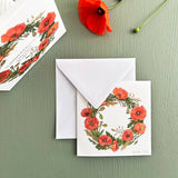 A charming square card folder from Koustrup & Cos Card Pack: Summer Wreaths, featuring 8 unique designs of poppy flower wreaths, adorns an envelope. Nearby, a vibrant red poppy on green captures the essence of its elegant Danish design.