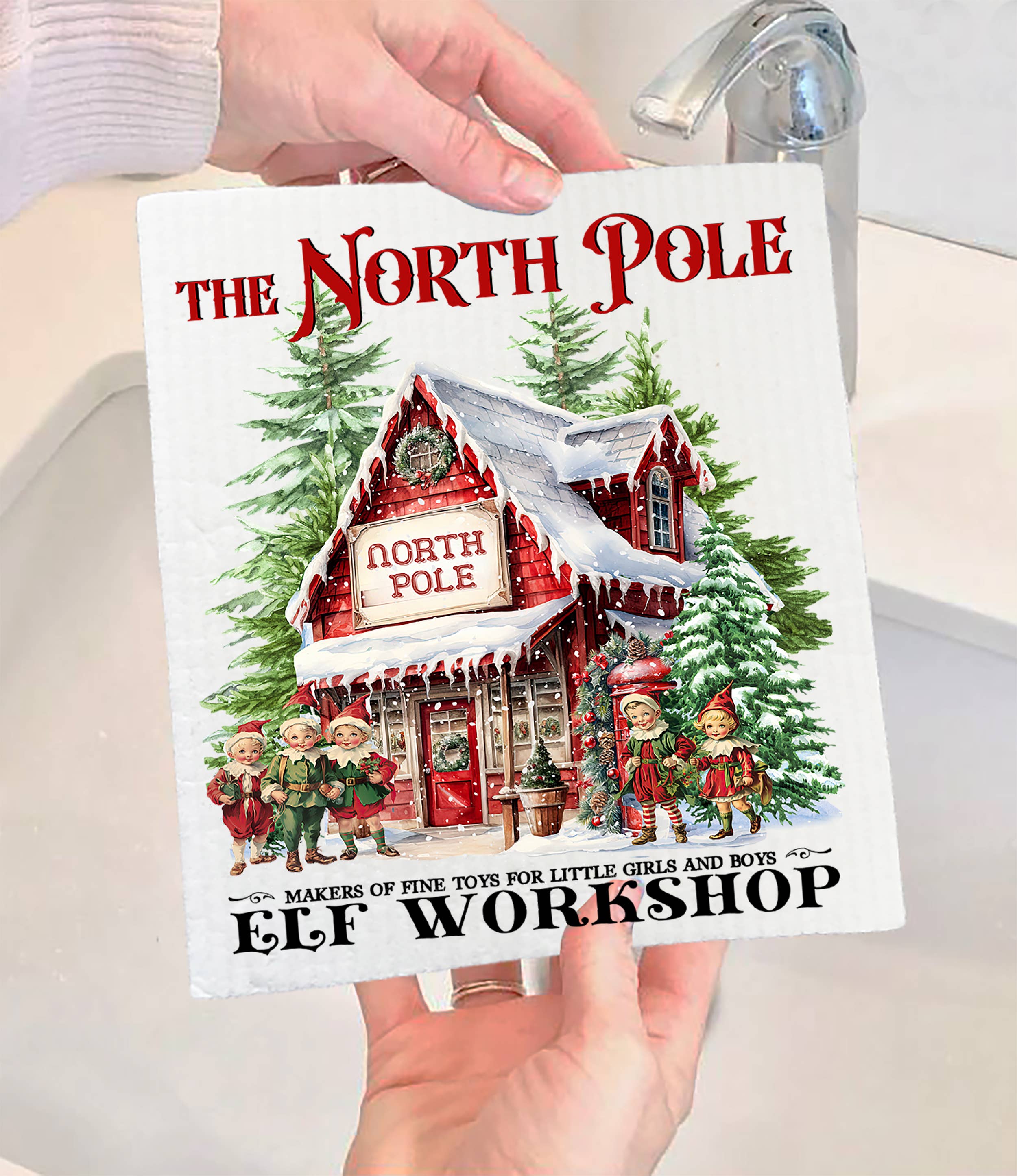 A person holds the Dish Cloth: Christmas Vintage North Pole Elfs Swedish Dishcloth, which features a festive design with elves, Christmas trees, a North Pole workshop, and "The North Pole Elf Workshop" text.