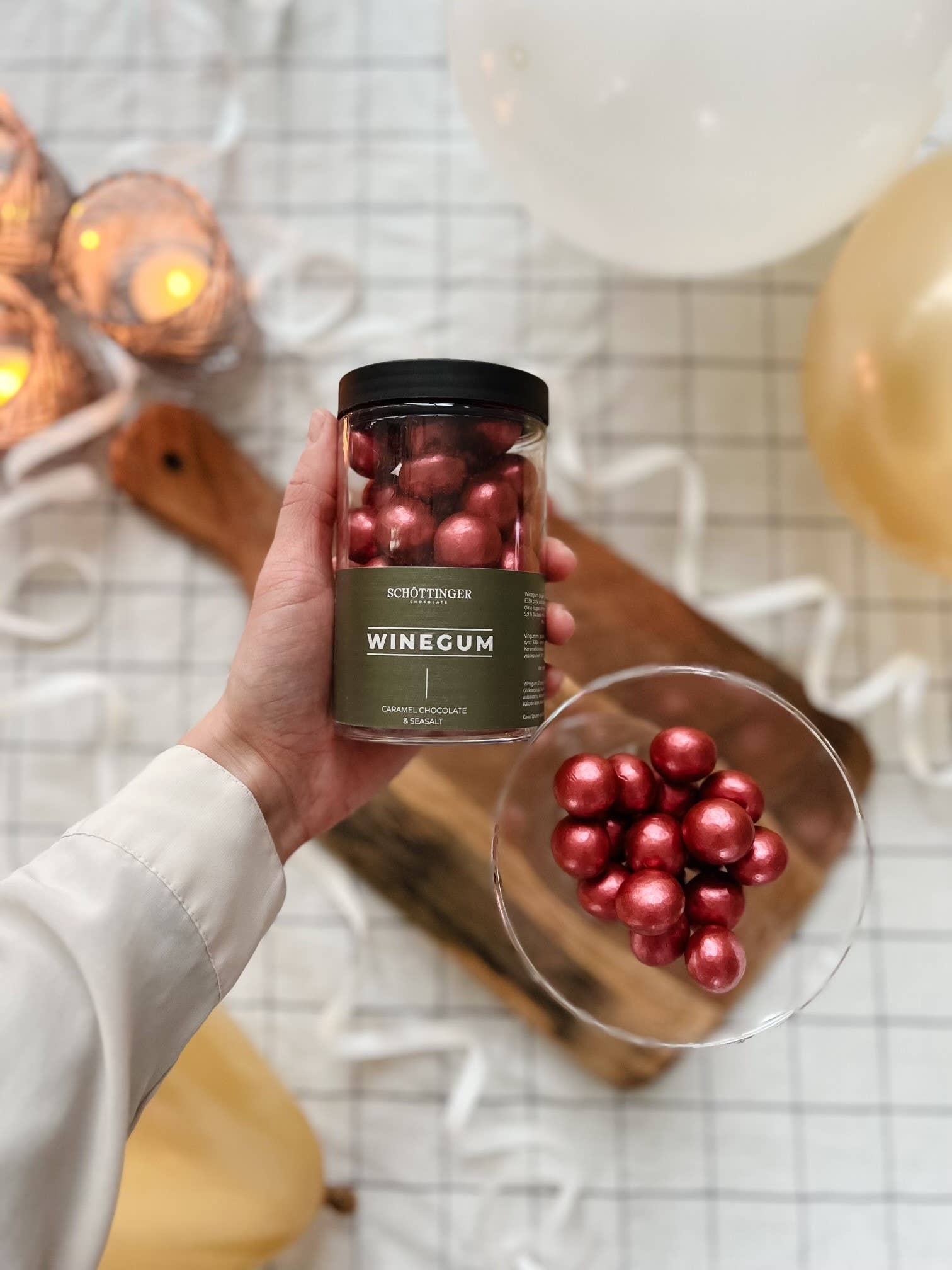 A hand holds a jar of Schöttinger Candy: Classic Winegum Seasalt & Caramel Chocolate 250 g over a glass bowl filled with red winegum balls, shining like red amber. Candles and balloons create a festive backdrop on the checkered surface.