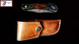 The Damascus Steel Pocket knife with a walnut wood handle and metal accents is displayed above a brown leather sheath on a black background.