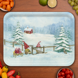 Birchwood tray with snowy scene of a red cabin, pine trees, and three gnomes skiing with gifts on a sled through a wooden gate; tray sits on table with fruits.