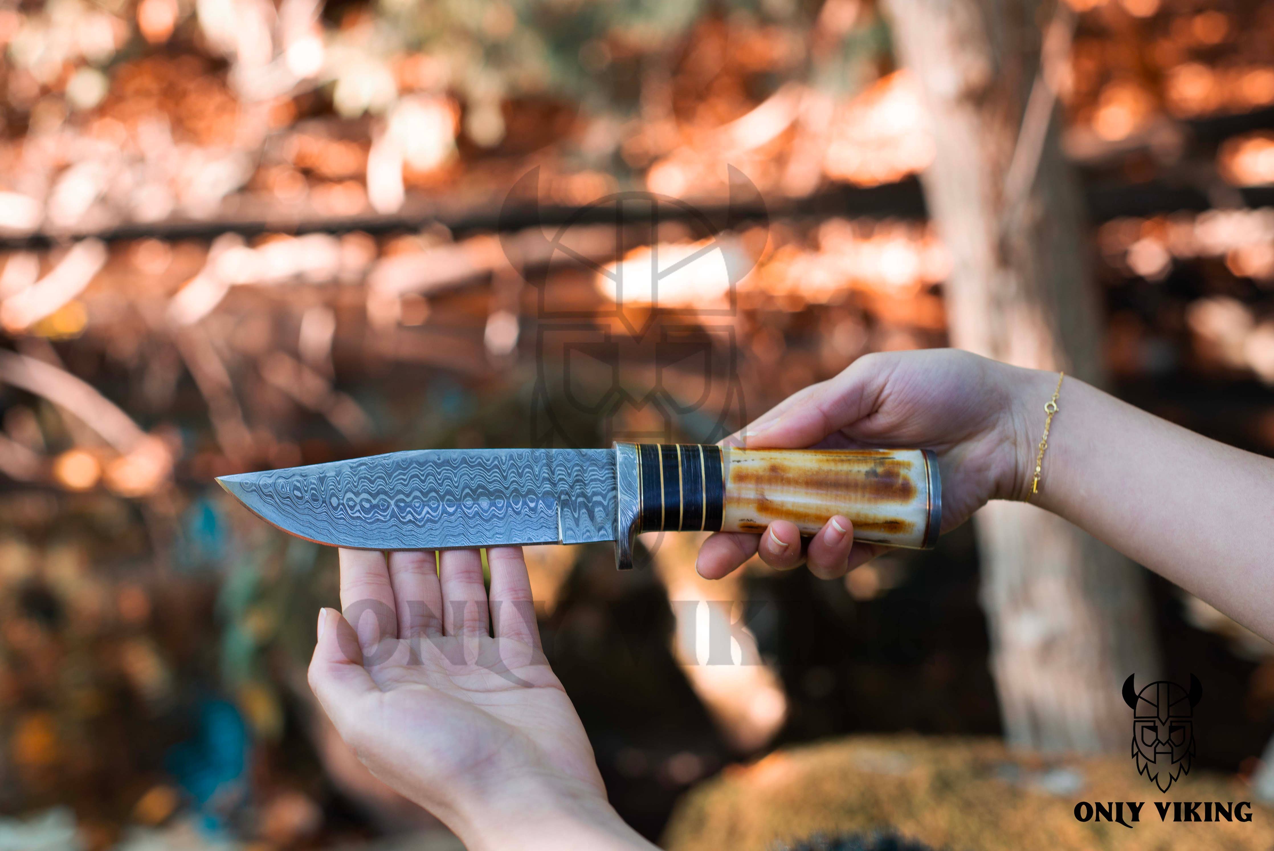 Hands holding the Knife: Damascus Hunting Knife With Locking Leather Sheath, showcasing its patterned blade and decorative handle against a blurred natural background, ideal for outdoor activities.