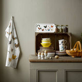 A kitchen counter features stacked dishes, yellow bowls, eggs, and a wooden basket. A wall-mounted rack holds a chicken-patterned towel. A wooden crate showcases the Colorful Hens on Birch Veneer serving tray and containers, adding charm to the scene.