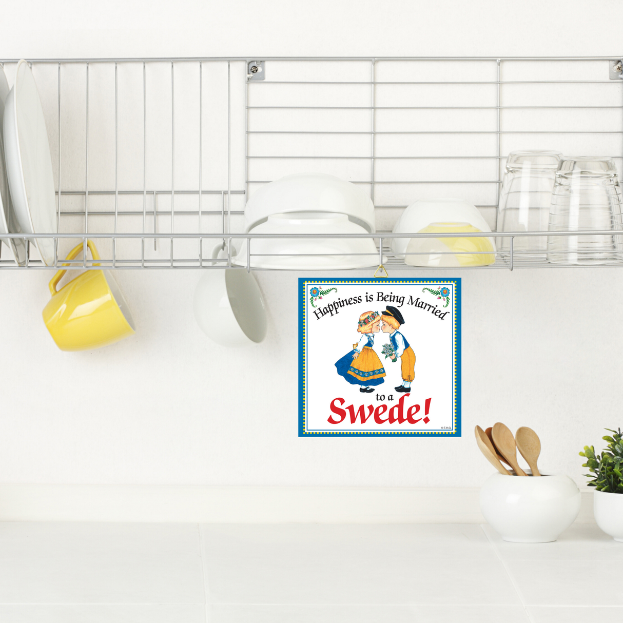 A kitchen shelf with dishes and mugs above a counter, featuring the 6x6 ceramic wall plaque Happiness is Being Married to a Swede with a couple in traditional attire.