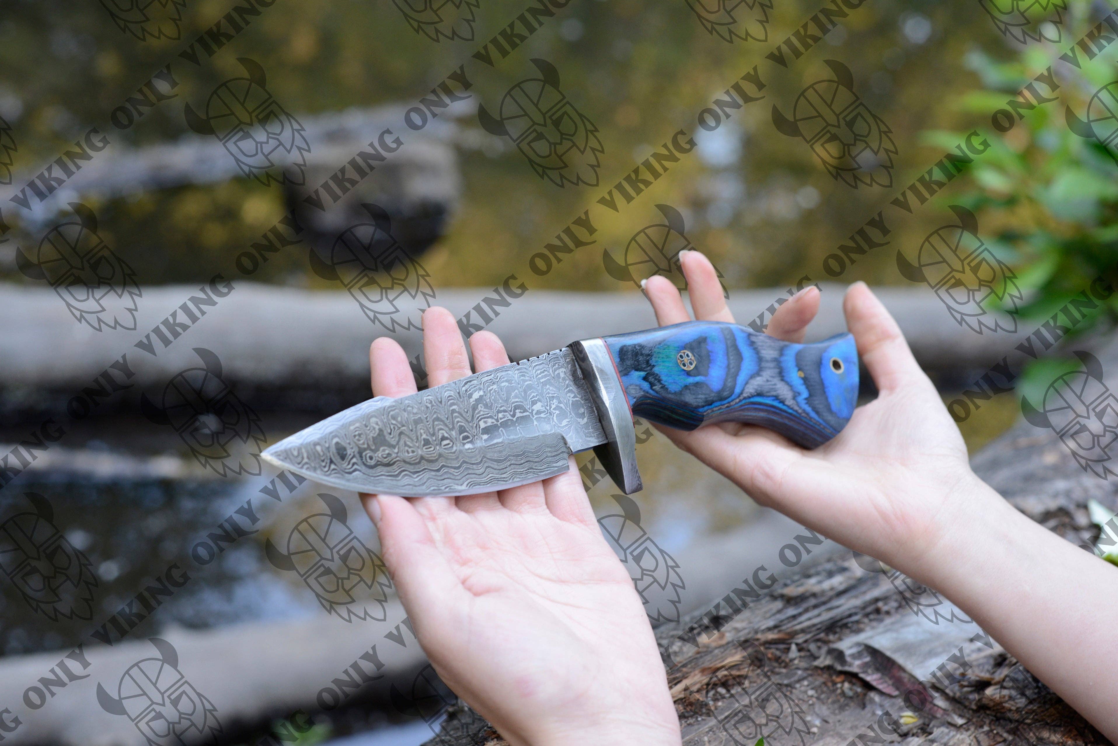 A person holds a large, patterned Premium Quality Damascus Utility Knife with a blue and black handle near a stream in the forest.