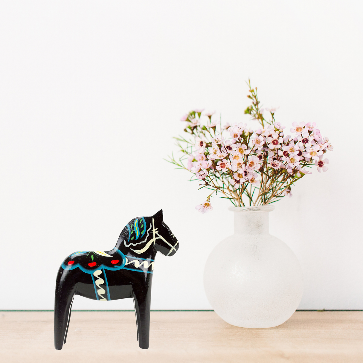 Black Dala horse figurine with colorful painted patterns next to a frosted glass vase holding pink flowers on a light wooden surface, white background.