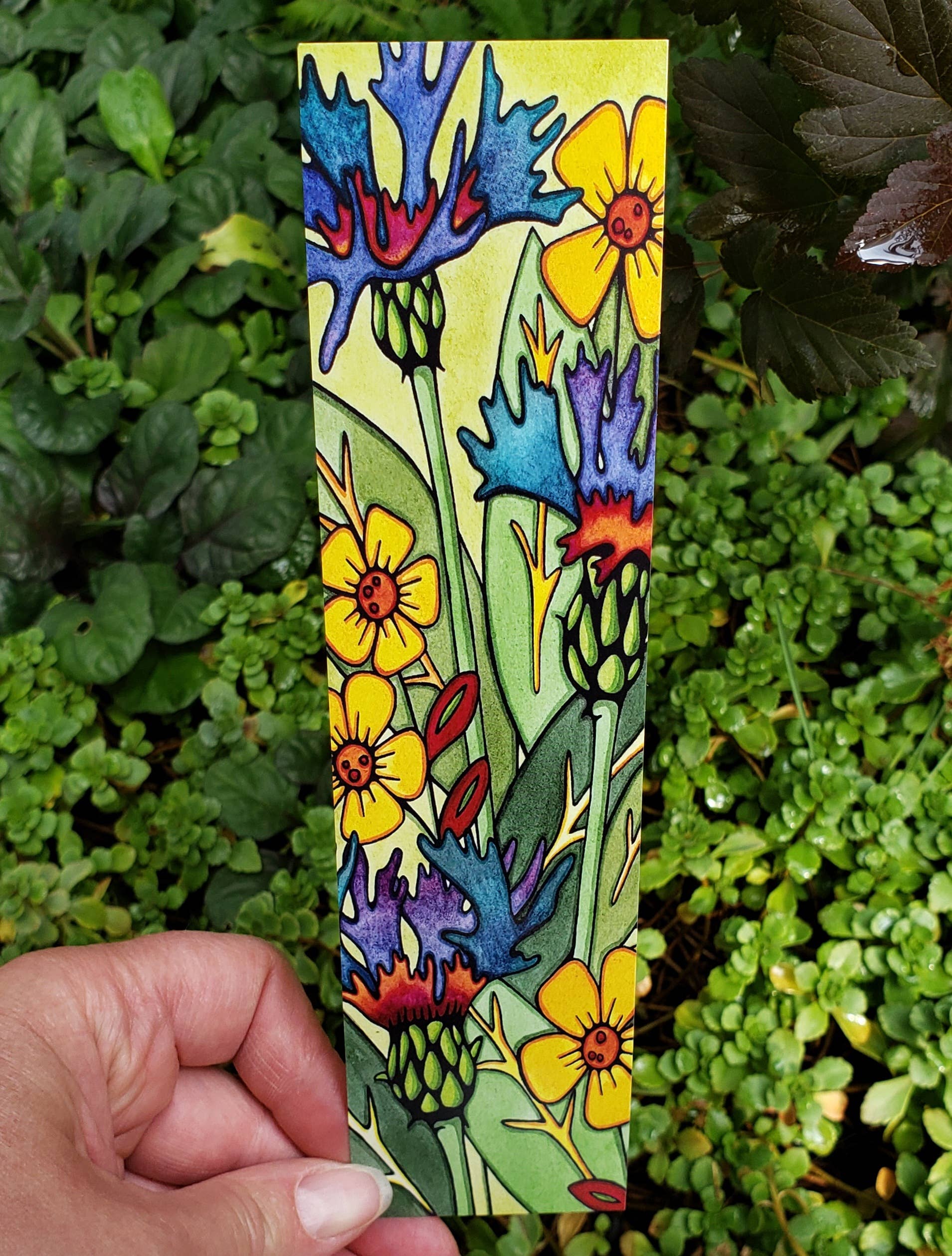 A hand holds the vibrant Mountain Bluet Bookmark from Sarah Angst Art, featuring colorful floral and leaf illustrations against green foliage. Proudly printed in the USA.