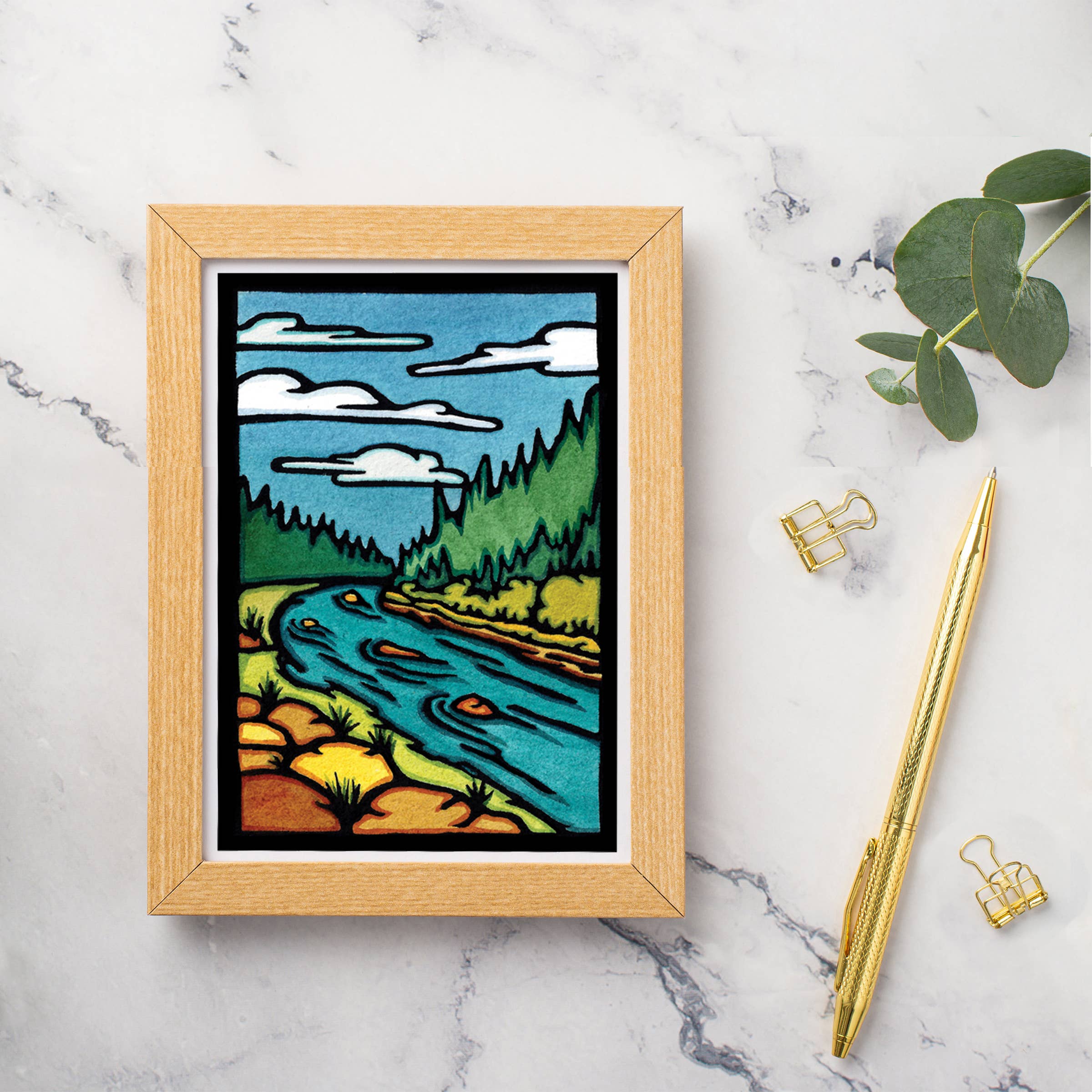 Greeting card featuring a landscape of a river flowing through a forest with clouds above, styled in the spirit of Linocut Prints. The card sits elegantly on a marble surface alongside a pen, clips, and green leaves, capturing the timeless elegance characteristic of Sarah Angst Art.