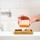 A hand places a bar of soap on a bamboo soap lift next to a bathtub with water running from the faucet, set against a white tiled wall.