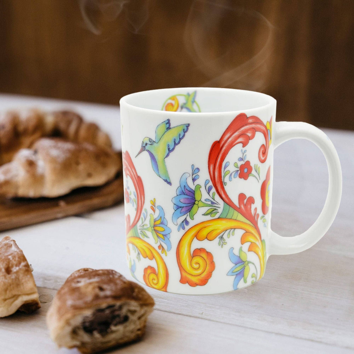 A 12 oz Colorful Norwegian Rosemaling Coffee Mug sits on a table near pastries, featuring vibrant floral and hummingbird designs with steam rising from the mug.