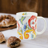A 12 oz Colorful Norwegian Rosemaling Coffee Mug sits on a table near pastries, featuring vibrant floral and hummingbird designs with steam rising from the mug.
