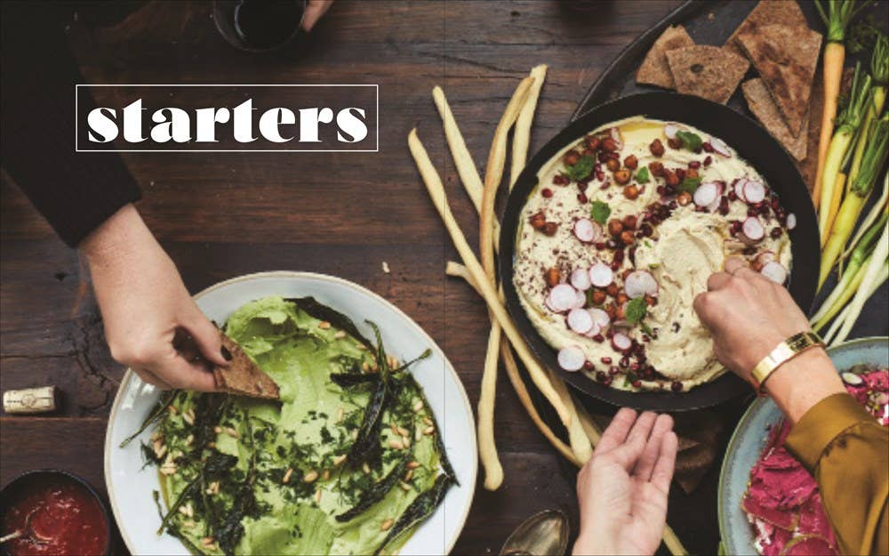 A gathering of modern culinary delights showcases a vibrant spread of starters like topped hummus, a fresh salad, breadsticks, and pita. Three individuals eagerly dive into the offerings beneath the title "Starters" at the top left corner. This is a scene drawn from the book "Potluck.