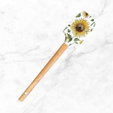 Wooden-handled spatula with a yellow sunflower-shaped silicone head featuring green leaves, resting on a white marble surface.