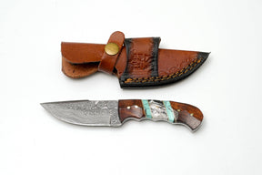 The Titan International Hand-Forged Damascus Steel Field Knife features a patterned blade and a custom hybrid wood & turquoise handle, shown beside a stitched, embossed brown leather sheath on a white background.