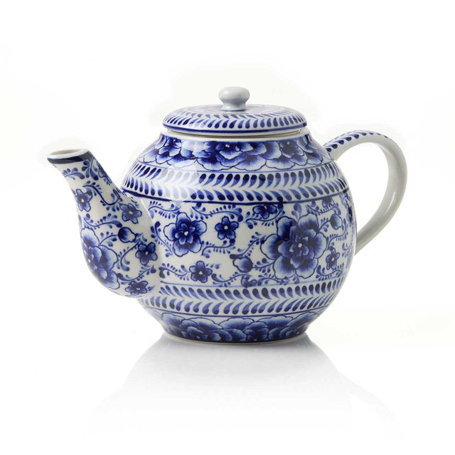 The Indigo Bloom Tea Infuser Teapot is a white ceramic teapot with a blue hand-painted floral design, curved spout, round lid, and handle, shown against a white background.