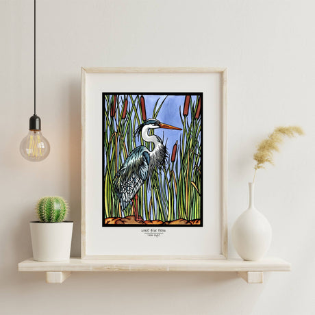 Blue Heron 8x10 Print by Sarah Angst displayed among cattails on a white wall shelf with a round plant, hanging bulb, and white vase with dried grass.