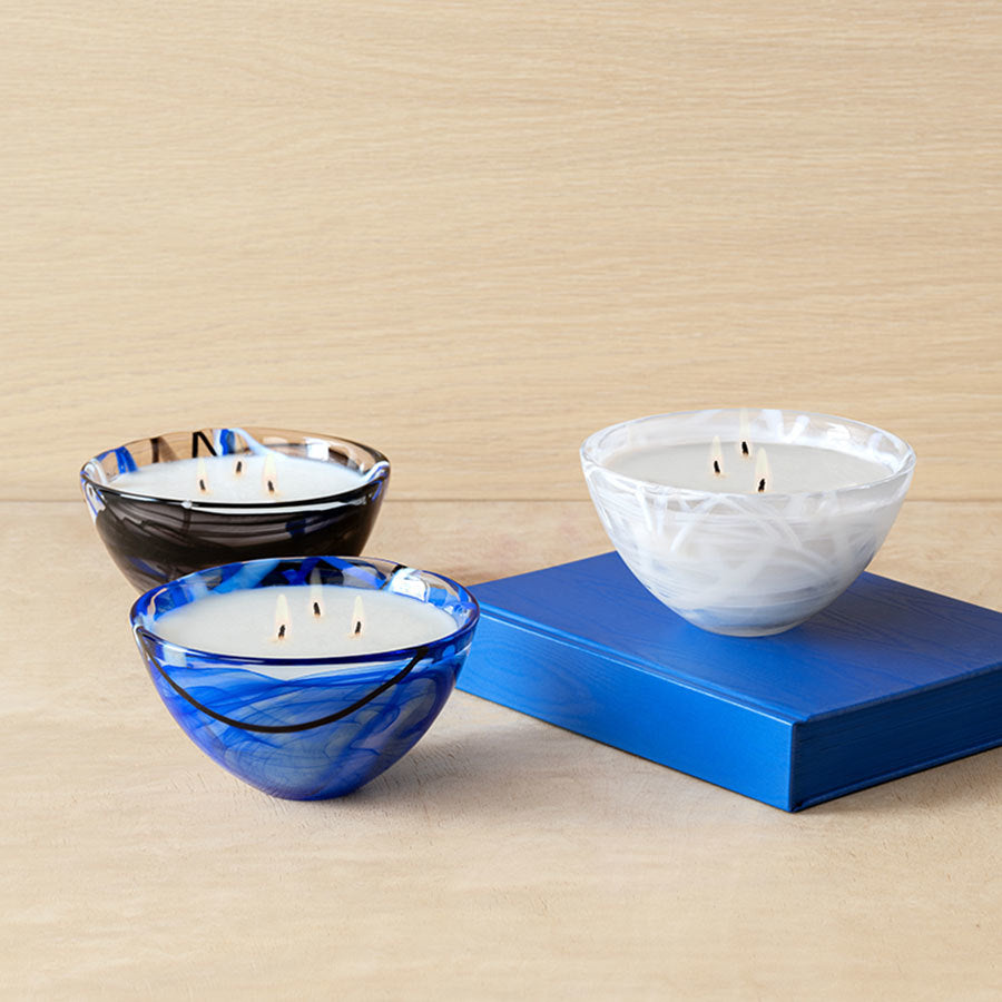 Three KostaBoda: Contrast Candle Blue decorative bowl candles featuring Coastal Bloom designs in blue, black, and white sit elegantly on a wooden surface next to a blue book, perfect for adding home fragrance to any room.